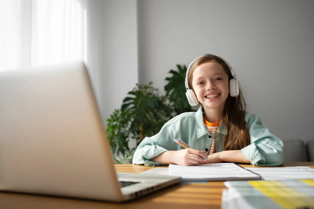 Smiling girl wearing headphones writing on a notebook with a laptop in front on the table