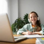 Smiling girl wearing headphones writing on a notebook with a laptop in front on the table