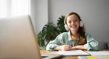 Smiling girl wearing headphones writing on a notebook with a laptop in front on the table