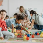 Children playing with train set and balls on floor with teachers in background with other children