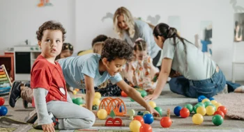 childcare-children-playing-pe Children playing with train set and balls on floor with teachers in background with other children