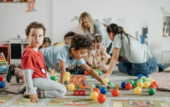 Children playing with train set and balls on floor with teachers in background with other children